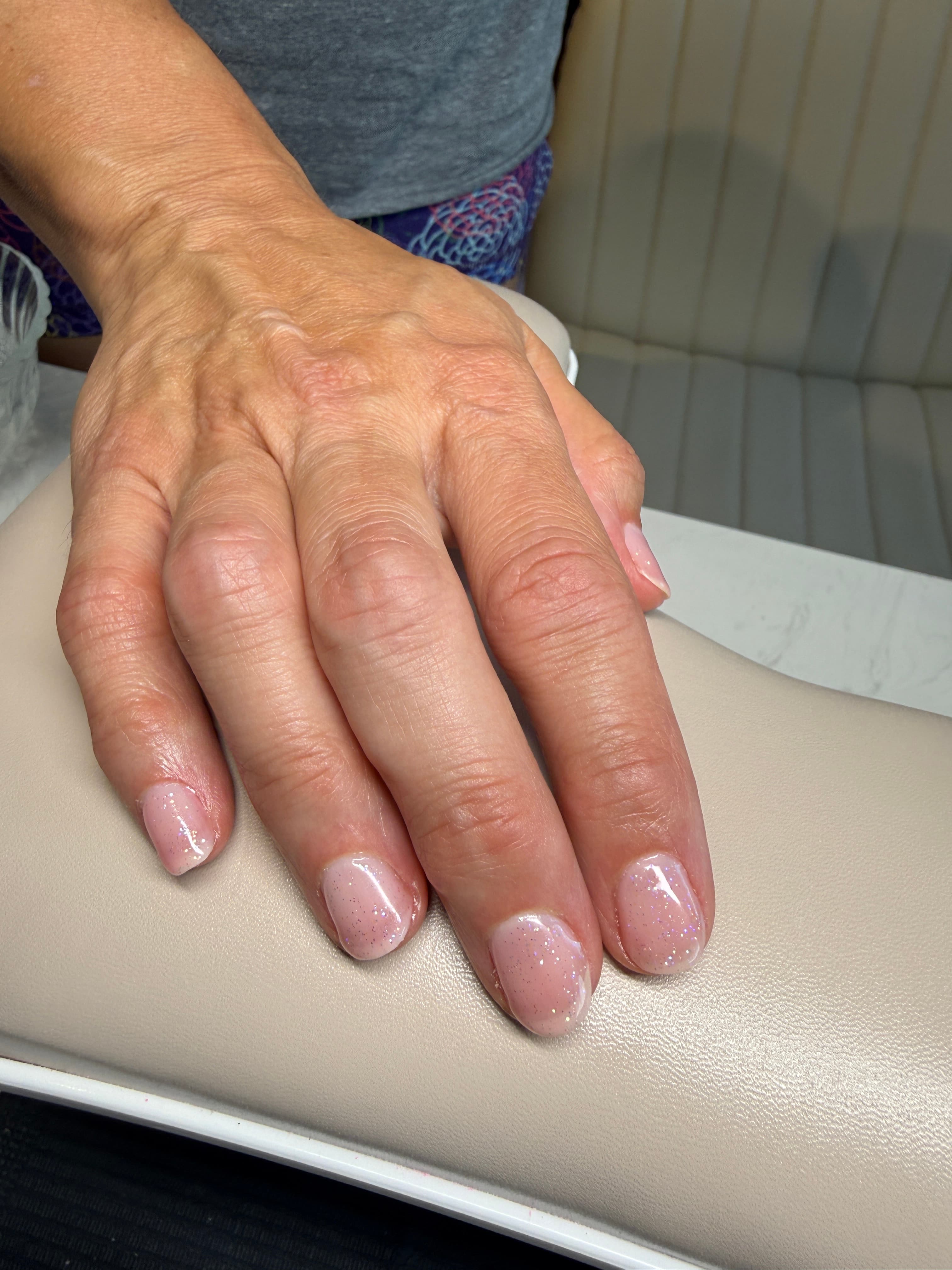 A close-up of a hand with glossy, glitter-flecked natural-pink gel nails resting on the cream leather of the studio's manicure chair.
